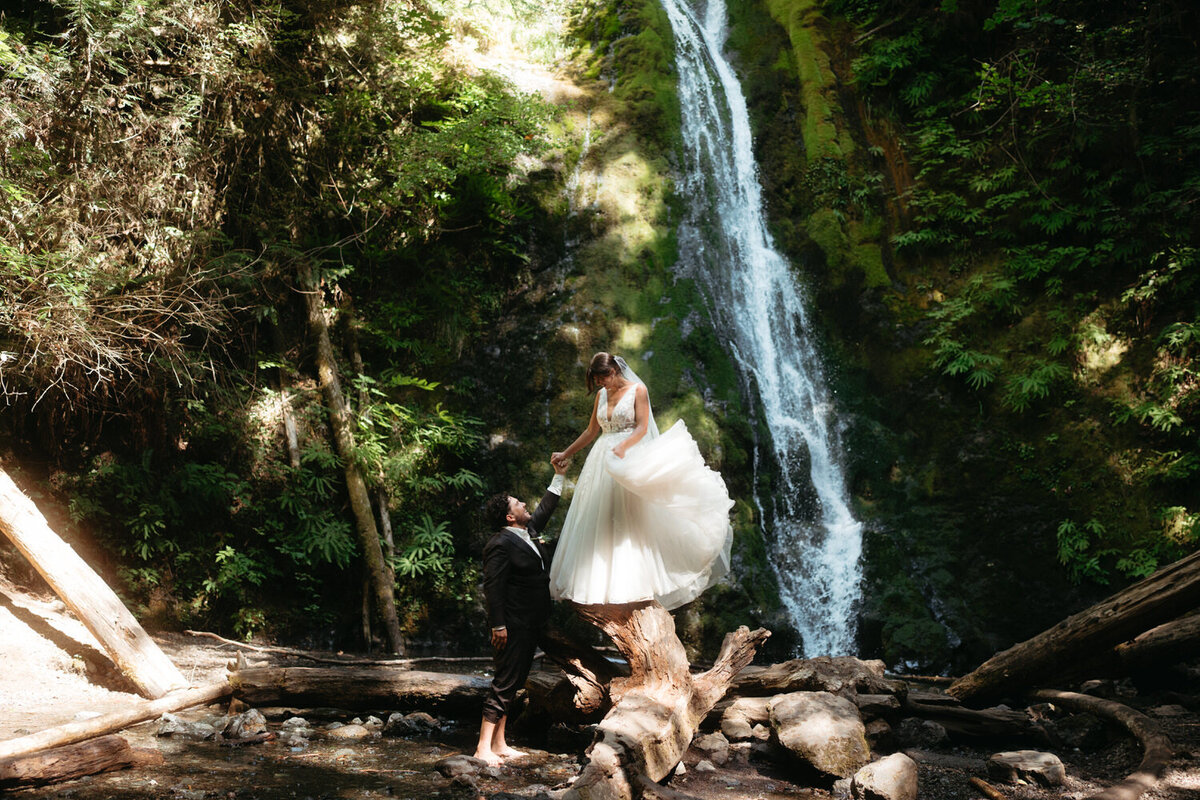 Olympic National Park wedding. Bride stands on a log looking down at groom. 