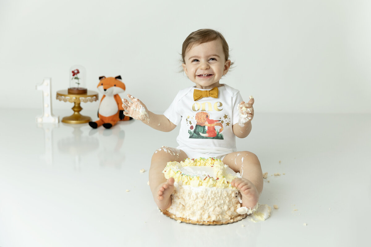 A young toddler sits in a cake wearing a gold bowtie and white shirt for his first birthday