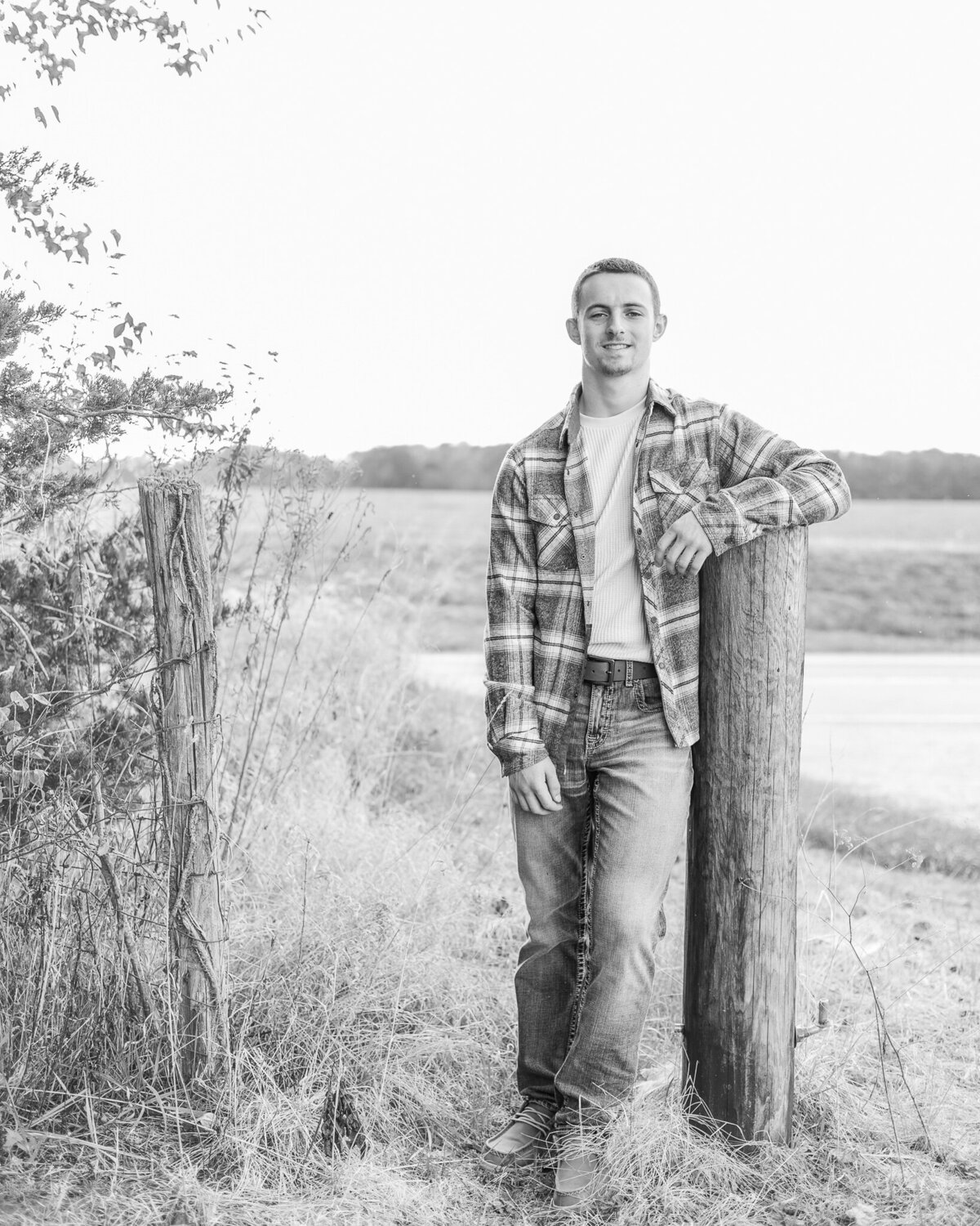 black and white photo of a high school senior male, leaning on a post, wearing jeans, flannel jacket, shirt, smiling at the camera