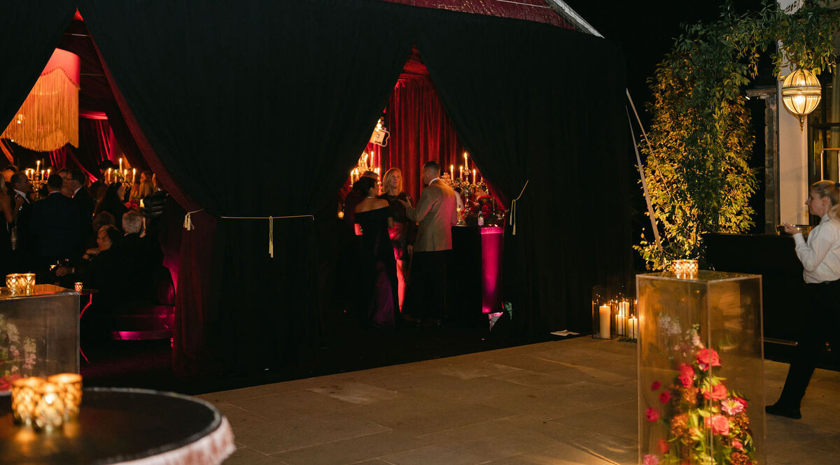 The information you provided has been updated to include Clayton Films as videographers at the event. Thank you for the addition of this detail. The photo was taken at a tented wedding in Virginia, captured by photographer Elisabetta Marzetti. People at an evening event gather inside an elegant, canopy-covered outdoor venue in this beautiful setting. Dark curtains and warm lighting enhance the atmosphere, creating an intimate and cozy space. In addition, candles, floral arrangements, as well as videographers from Clayton Films, have been tastefully incorporated to adorn the area, contributing to the festive ambiance. A waiter is also seen standing to the right, adding to the overall elegance of the scene.