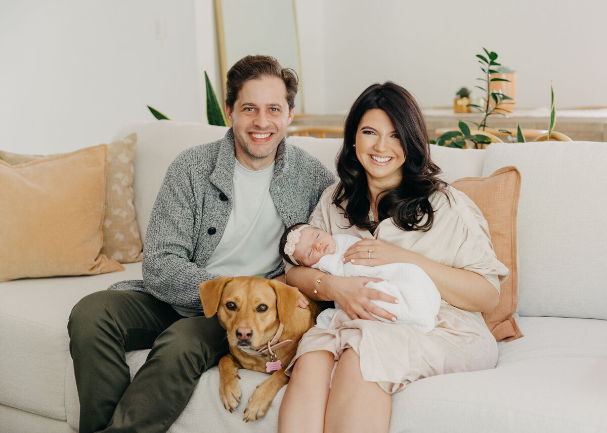 Family portrait of mom, dad, newborn baby, and dog sitting on the couch and smiling.