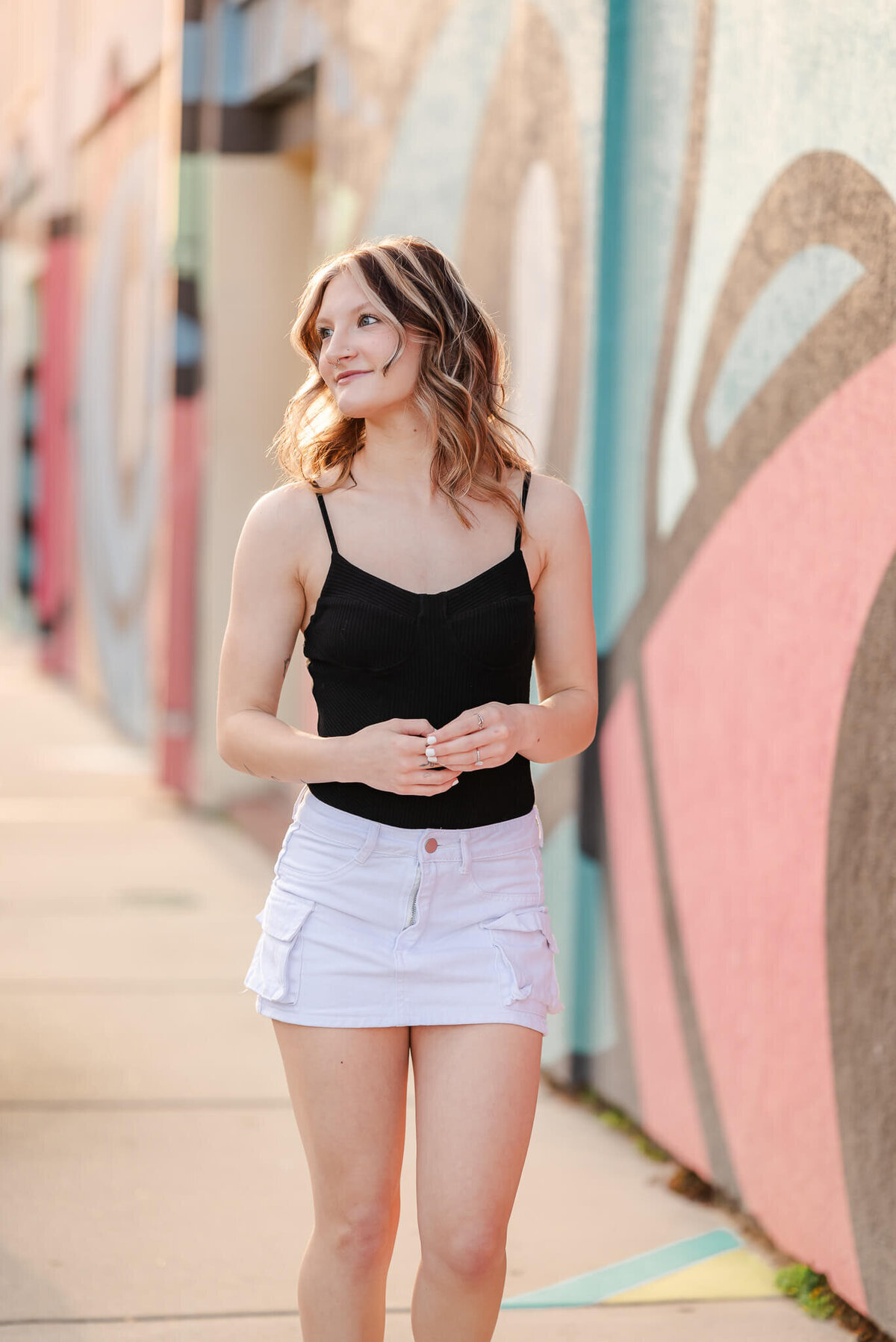 A high school senior walks down a sidewalk near a brightly painted mural. She is wearing a blank tank top and white denim skirt.