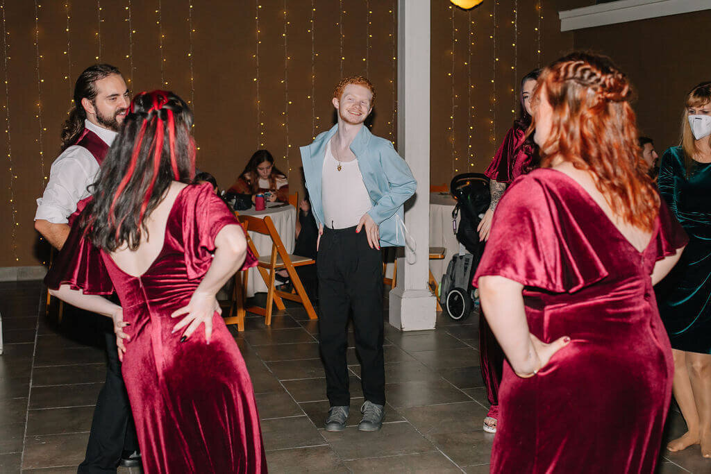 A group of people, including two women in matching red velvet dresses, dance together. String lights hang in the background, and other guests sit at tables, watching and chatting.