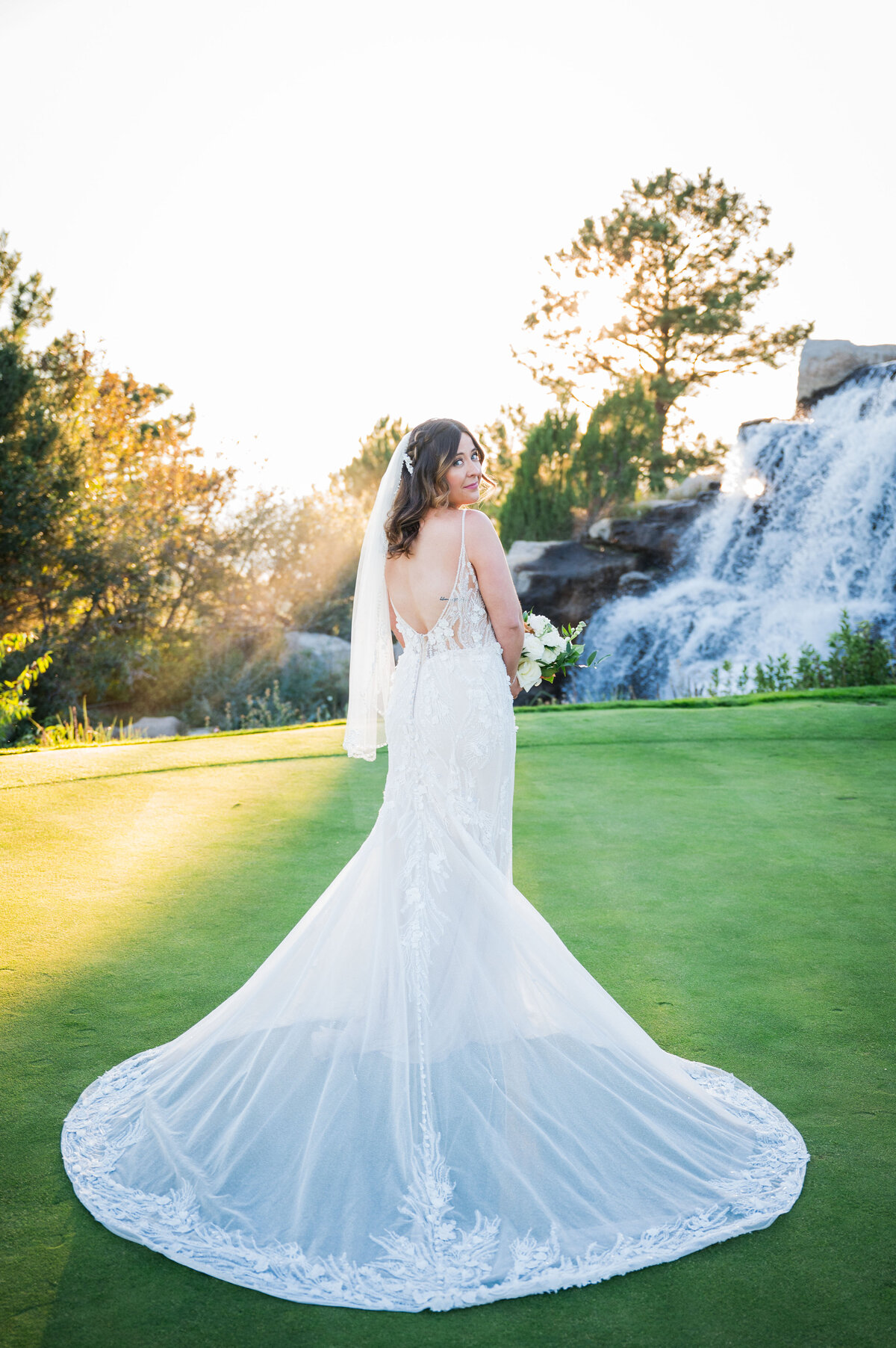 A bride looks over her shoulder at the camera with a golf course waterfall in the background.