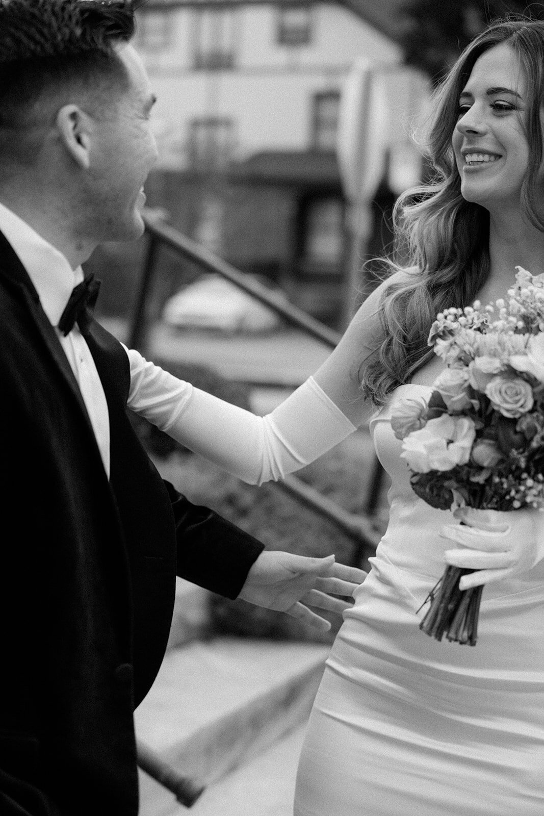 Groom in a black tux smiles at his bride in a white dress with white gloves while she smiles back on their elopement day. 