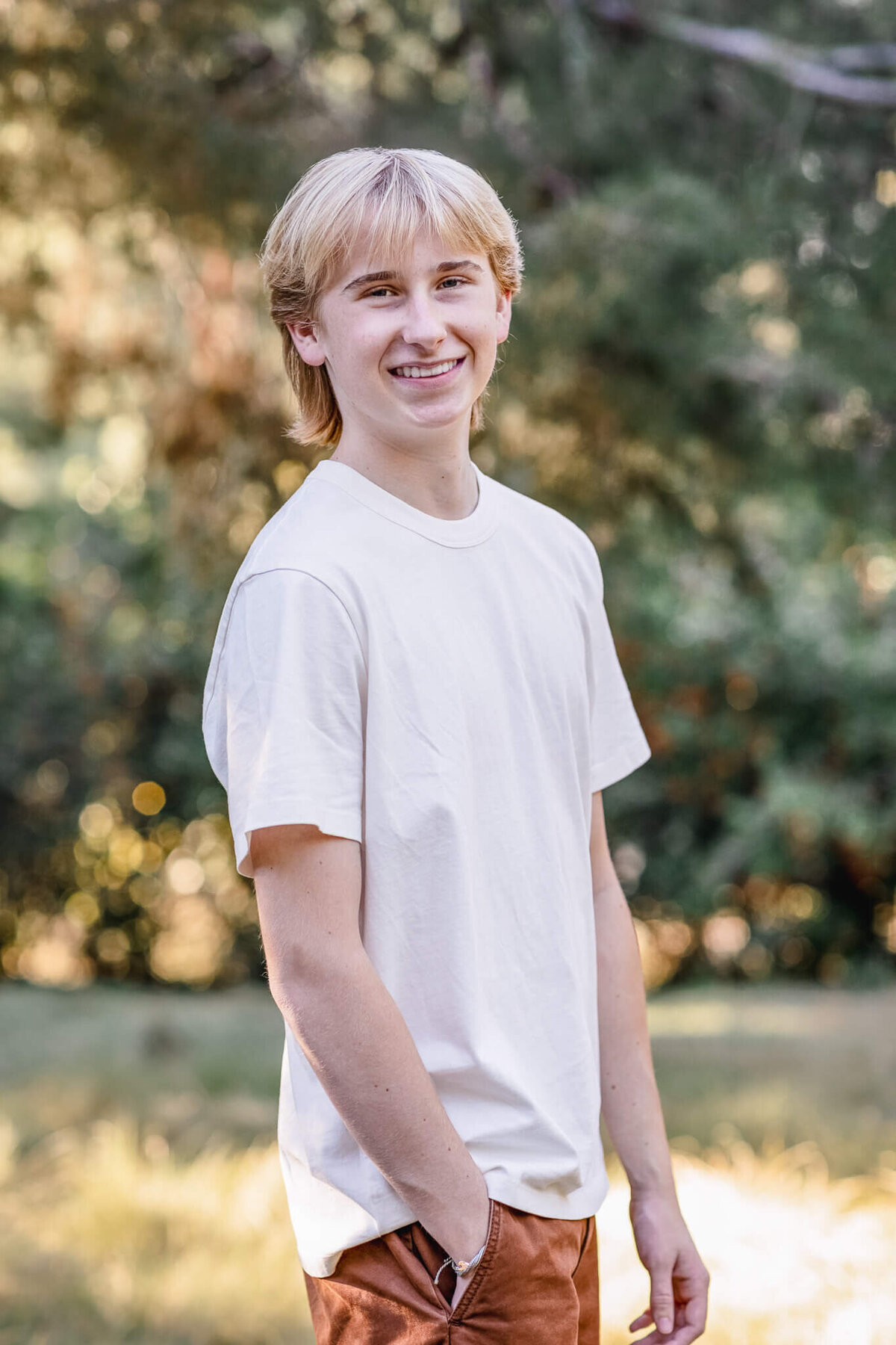 A teen boy with platinum blonde hair wearing a white t-shirt and brown pants with his hand in his pocket, looking past his right shoulder at the camera and smiling with greenery behind him.