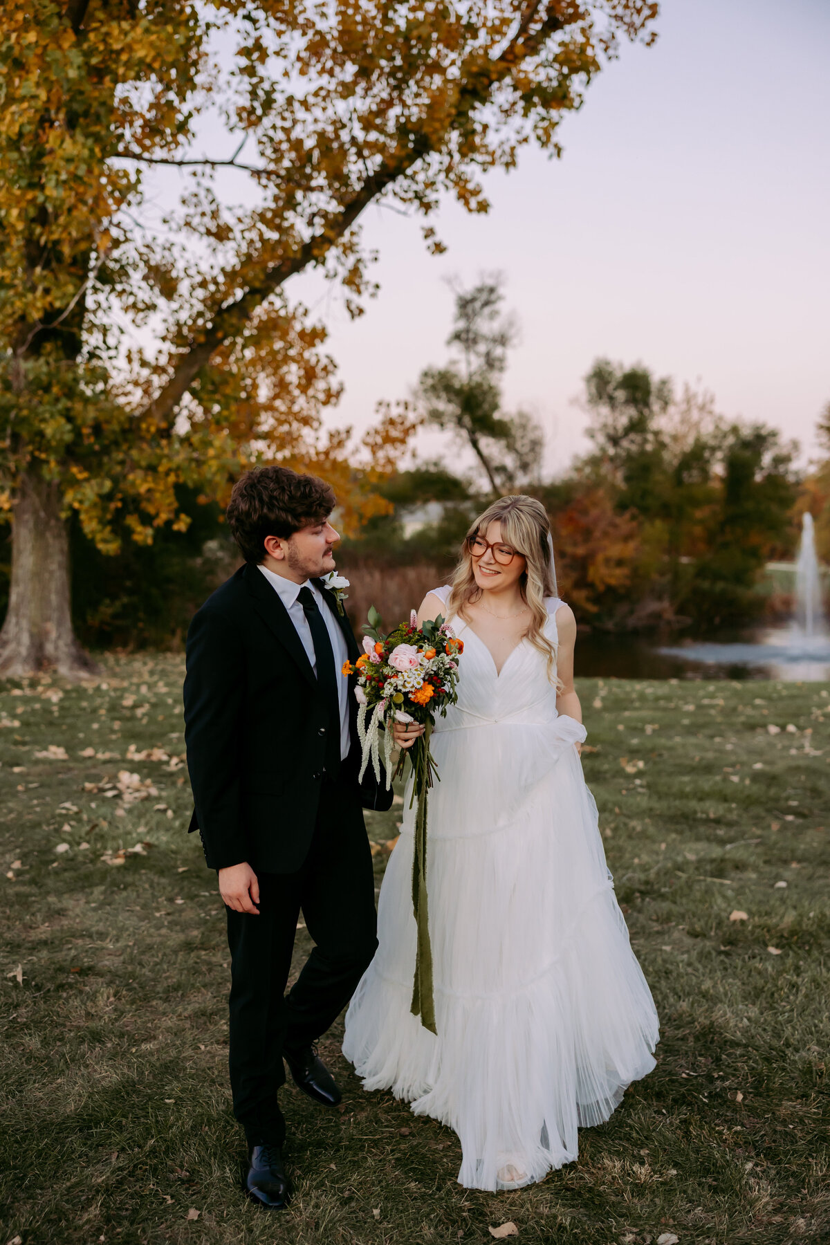 This photo shows two newlyweds walking in front of a pond with a fountain at Union 12 in Indiana. The groom is holding the brides bouquet.