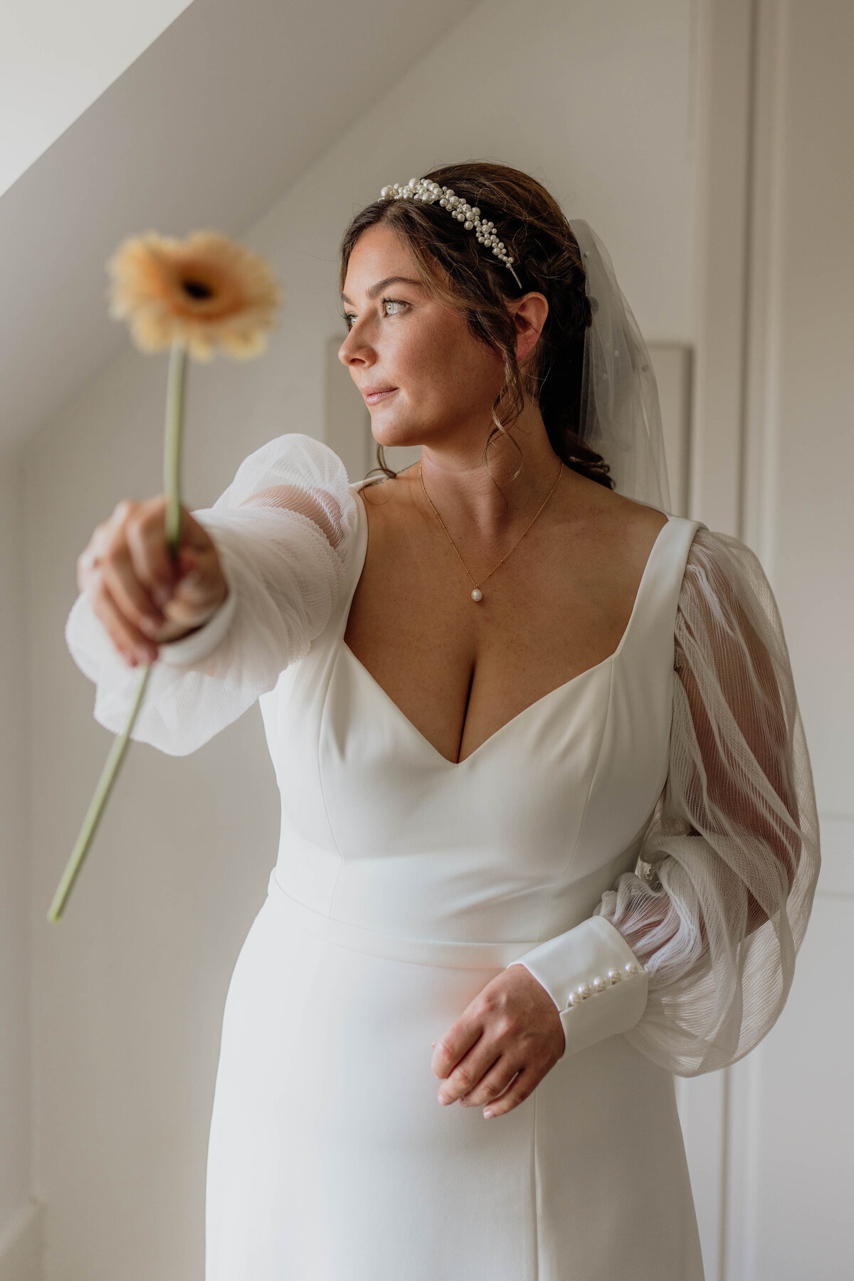 A bride wearing a white wedding dress holds an orange flower towards the camera.
