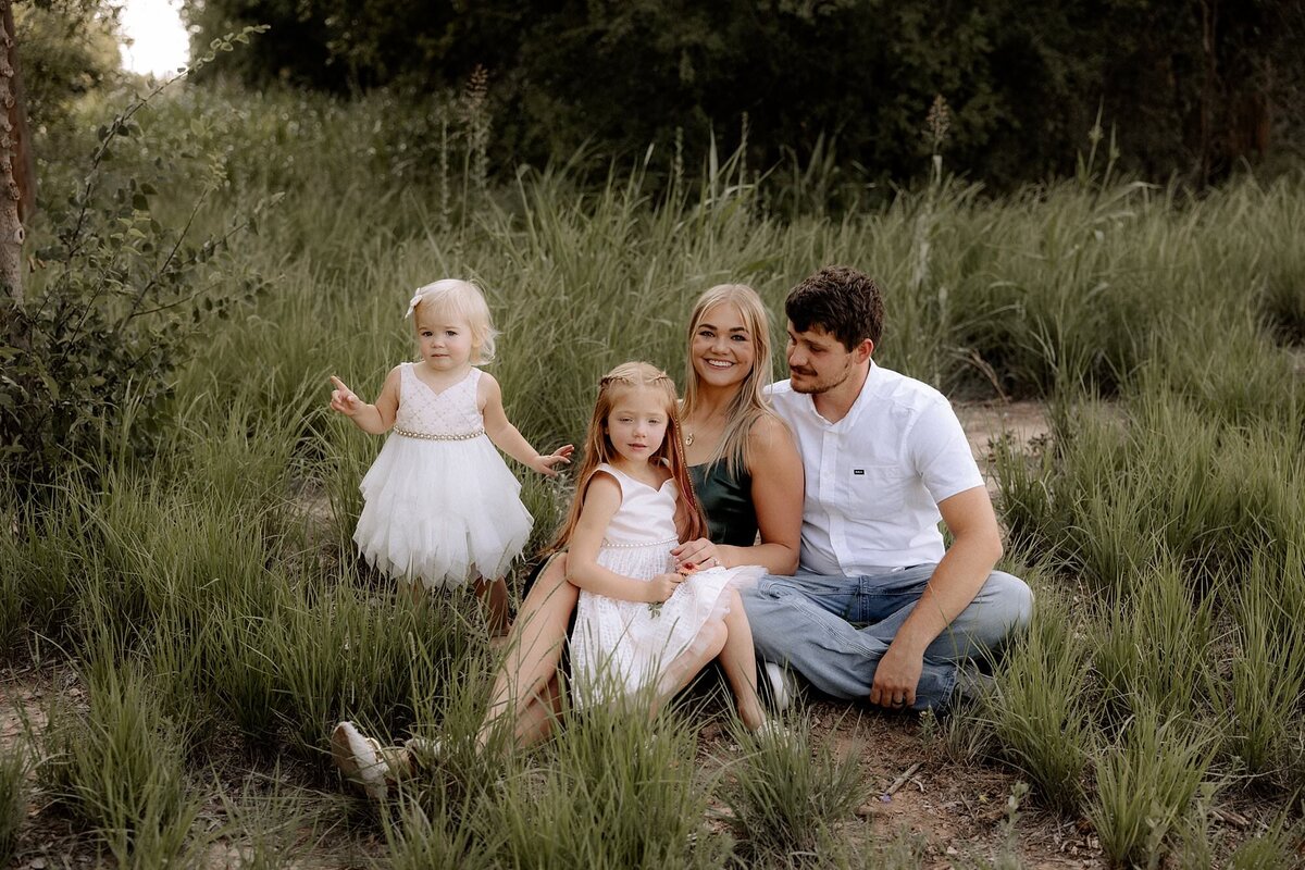 family of four sitting in a green field. The two girls are dressed in white summer dresses, mom is in a green emerald dress and dad is wearing a white button up shirt and demin jeans.