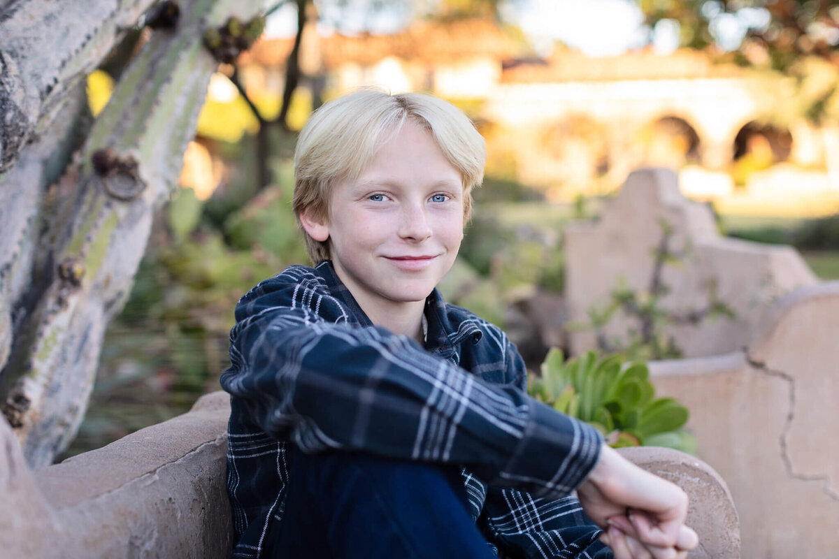 A close-up of a teen boy with blonde hair smiling at the camera with greenery and brick buildings behind him.