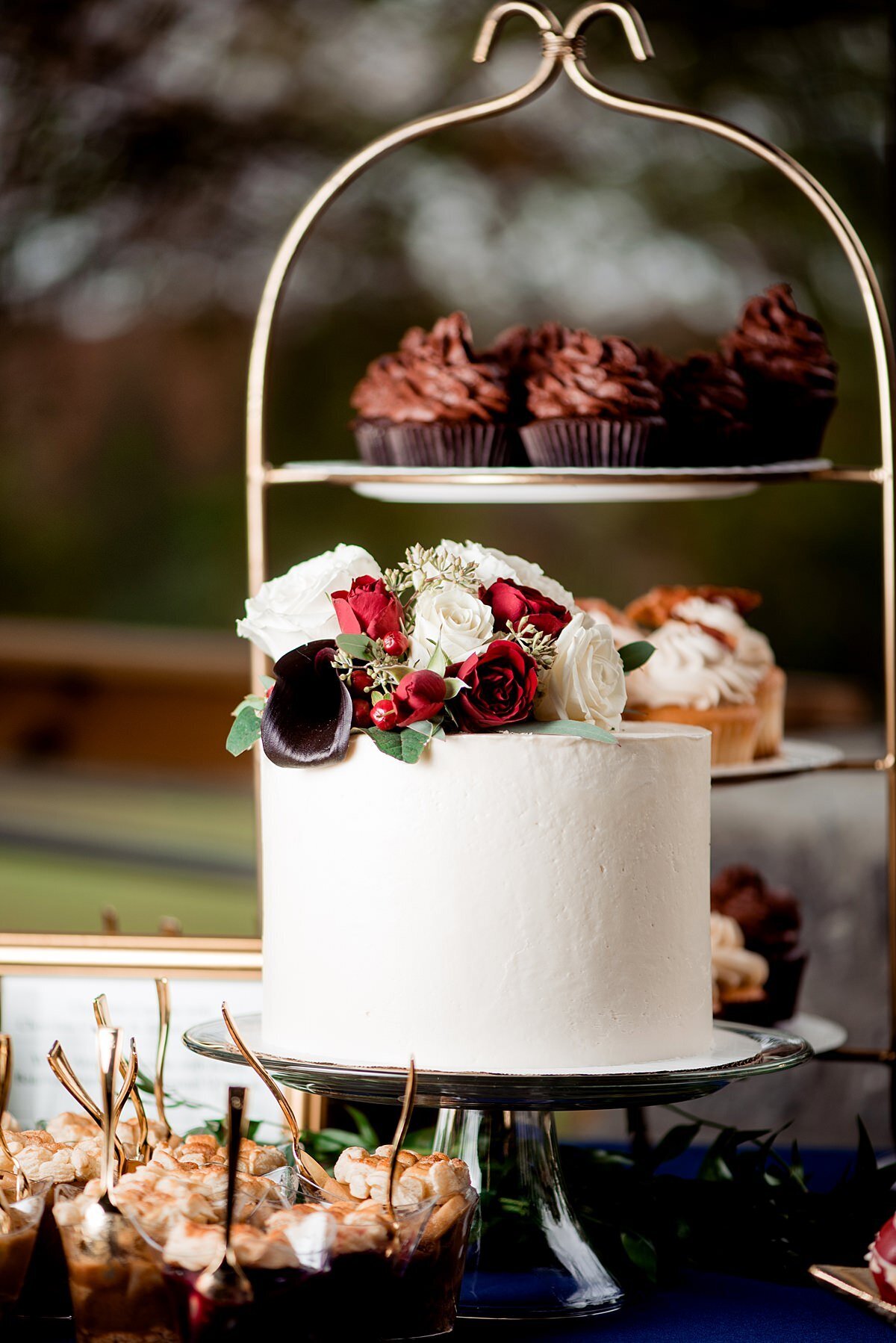 Small white wedding cake topped with white and red roses on a footed silver cake stand. Behind the cake is a three tier dessert stand with assorted cupcakes. In front of the cake are small dessert shooters.