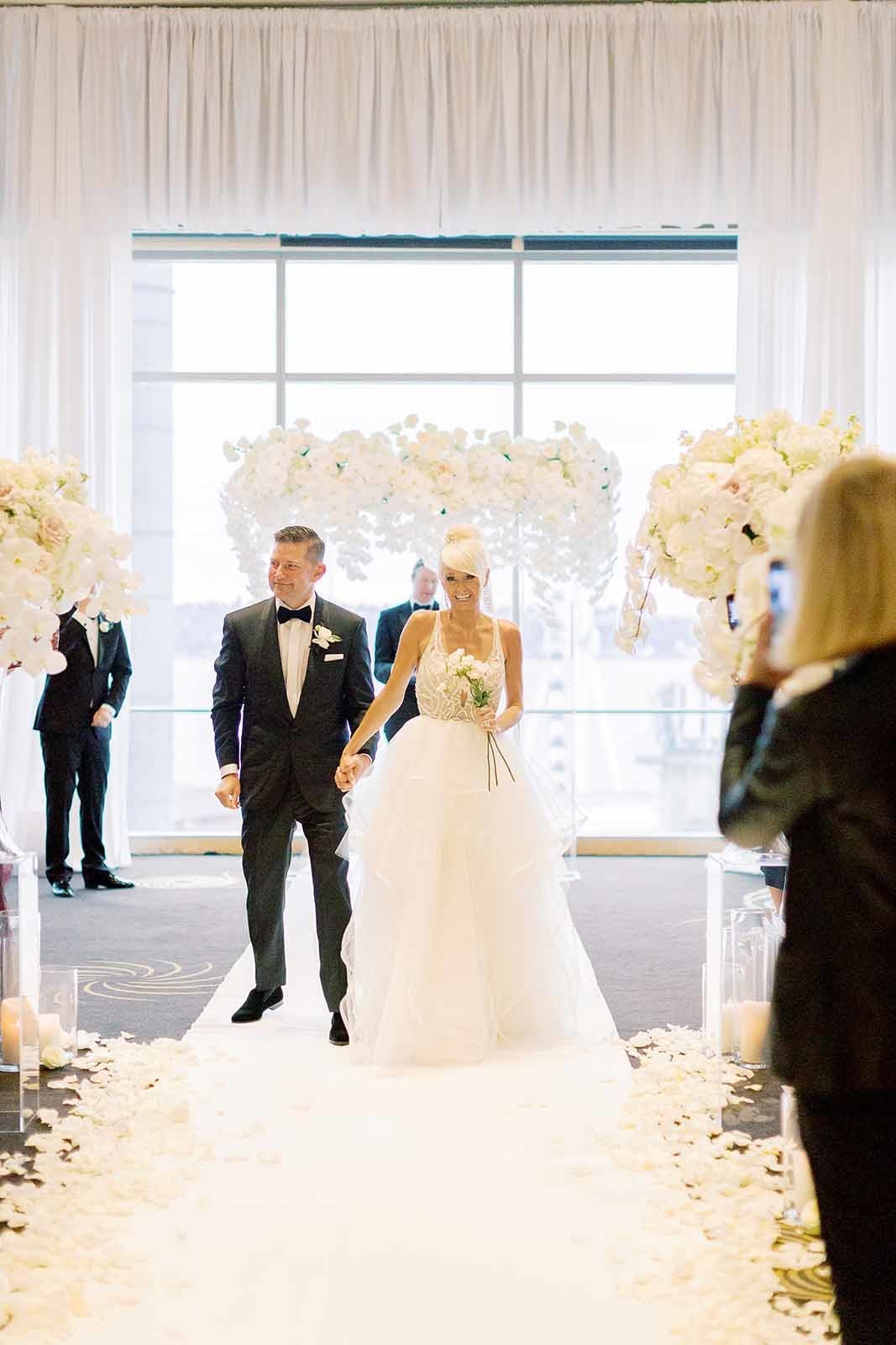 all white wedding ceremony and bride and groom walking down aisle with white orchid ceremony arch in back