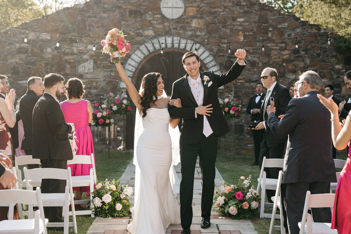 Bride and groom celebrating as they walk back up the aisle surrounded by cheering guests, with lush ground floral arrangements lining the ceremony path for a romantic floral-forward design.
