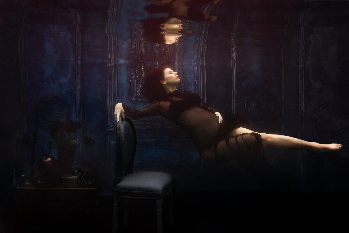 An ethereal underwater maternity photo showcasing a woman gracefully reclining against a vintage chair, her sheer black gown floating effortlessly in the dark water. Captured by a Dallas underwater pregnancy photographer, this dramatic scene is accentuated by rich textures and delicate reflections above, creating a hauntingly beautiful composition.