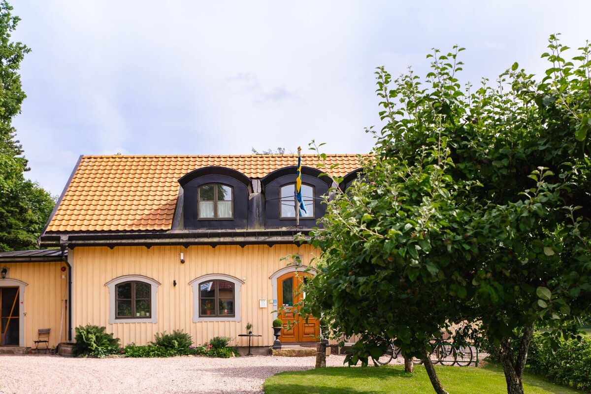 Framsidan av ett charmigt pensionat med svensk flagga, träpanel och grönskande trädgård.