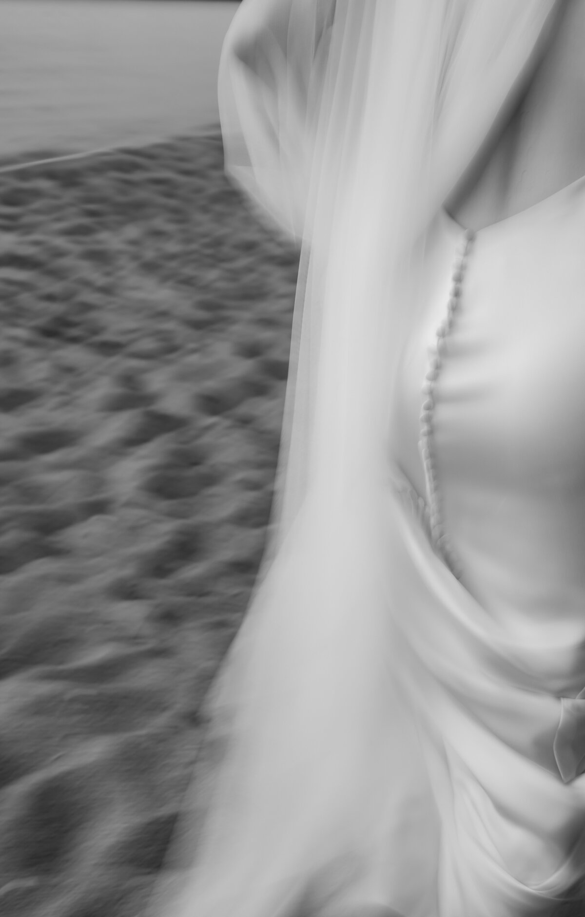 A blurred black and white image of a person in a flowing dress walking on a sandy beach captures the essence of elopement photography in New England. The focus is on the fabric's texture and smooth buttons, echoing the dynamic motion sought by a documentary photographer for New England weddings.