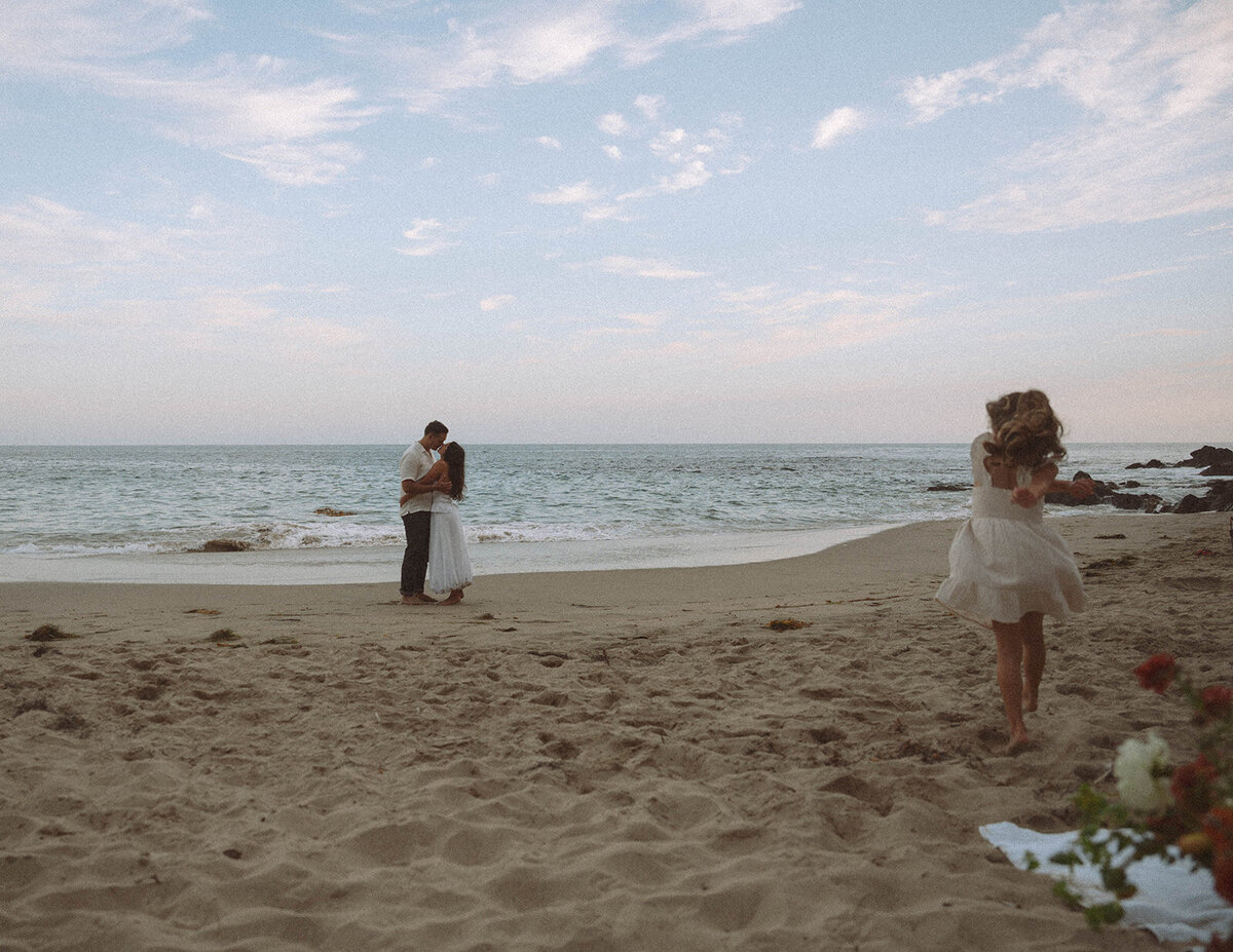 Parents embracing by the ocean at Laguna Beach while their daughter runs across the sand during a playful family photo session.