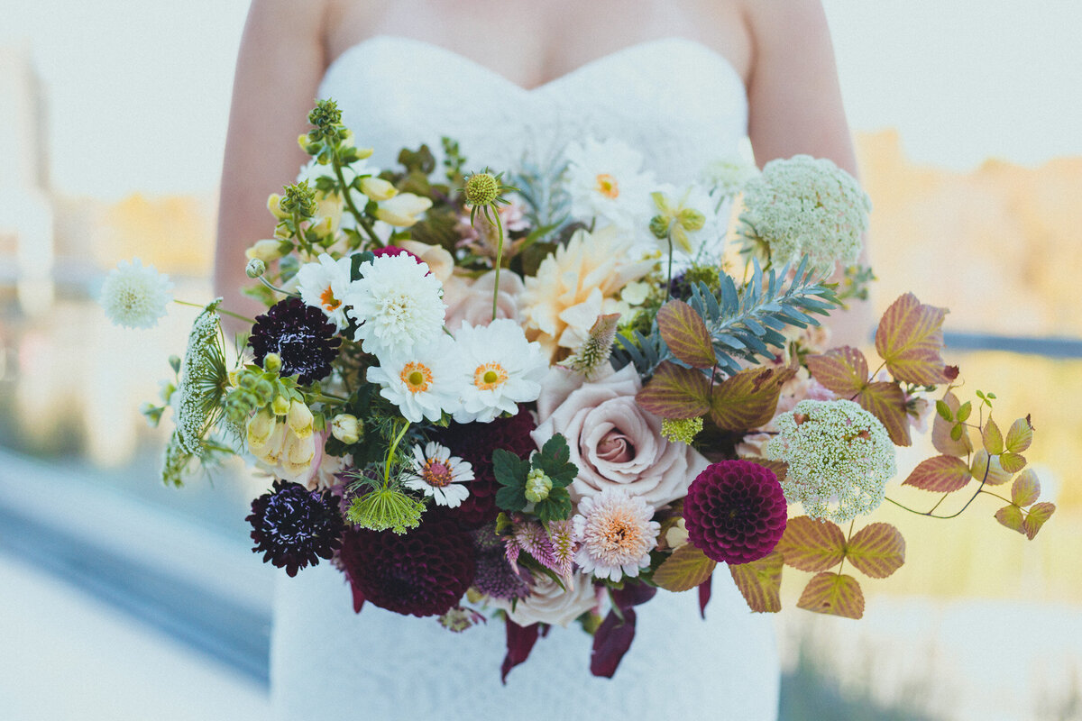 A bride holds an organic style bouquet of roses, dahlias, and cosmos, nestled in raspberry leaves.