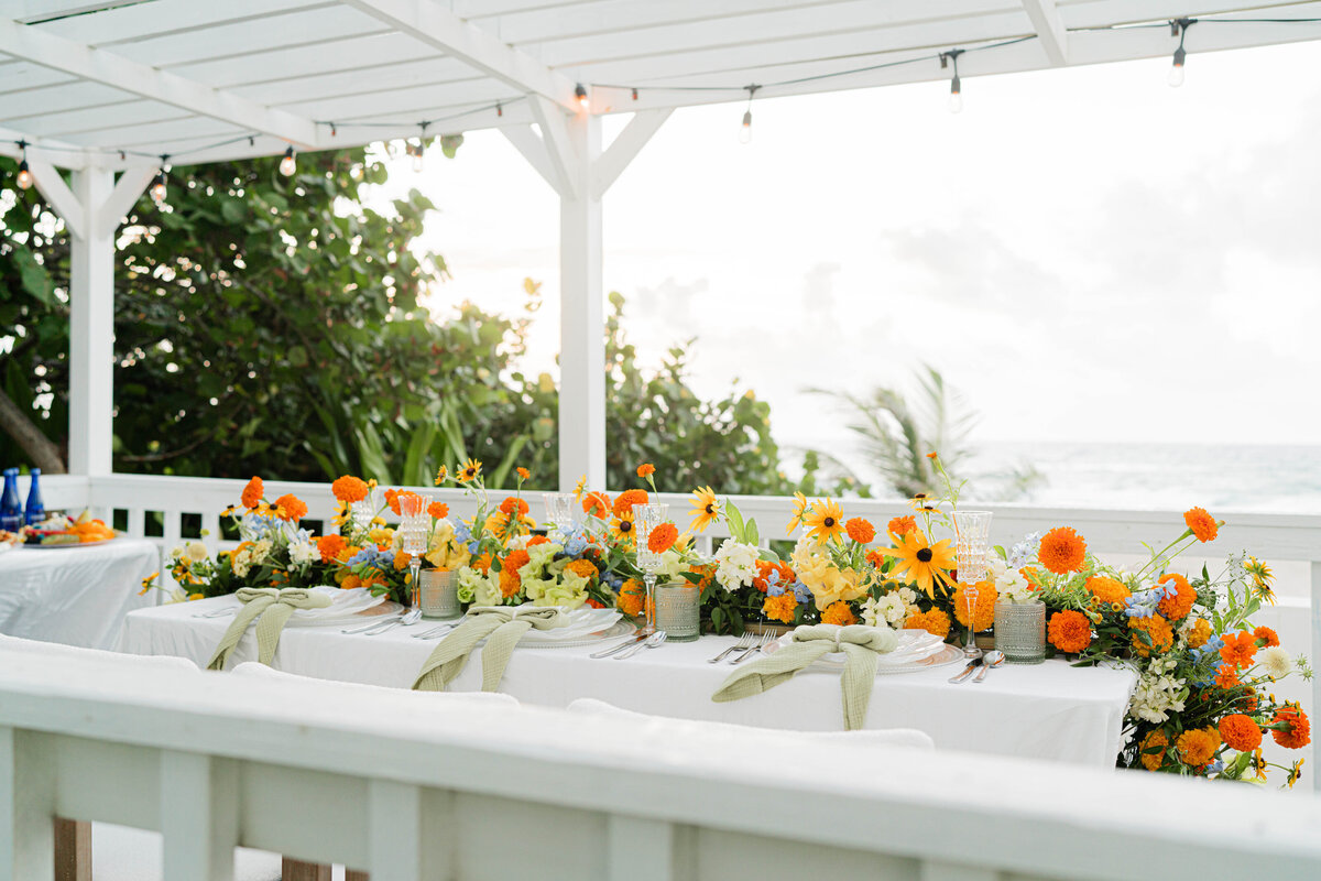 patio on the beach with a white on white palm leaf tablecloth and three iridescent seashell place settings adorned with a white, orange, blue and green floral table runner and green gauze napkin tied in a bow