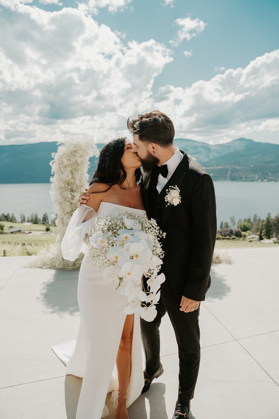 Kelowna Wedding Couple over looking the Okanagan Lake in Lake Country BC.  They are wearing modern sleek wedding attire with a gorgeous wedding dress and black tux.  A huge floral arch behind them filled with babysbreath.  A modern wedding day with a gorgeous look out!