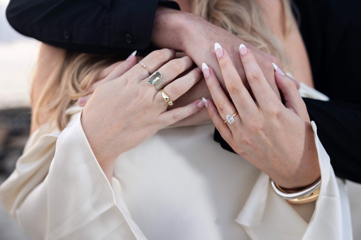 A close-up image of a bride-to-be’s hands gently holding her fiancé’s arm, showcasing her stunning engagement ring and elegant manicure. She wears multiple gold rings and a flowy white blouse, while his dark shirt contrasts beautifully. Captured by Heartful Films, a Jacksonville engagement photographer and videographer known for highlighting meaningful, intimate details with timeless style.