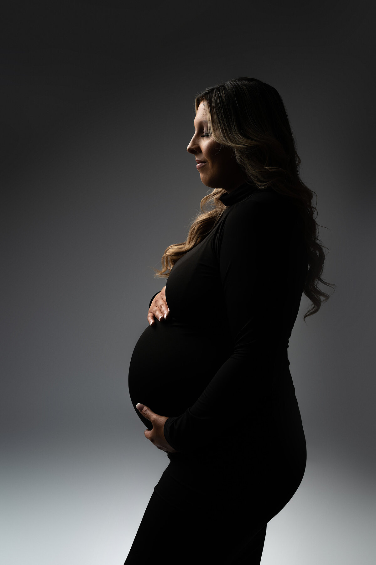 Elegant side profile of a pregnant woman in a black turtleneck, softly illuminated with moody directional lighting during a minimalist maternity session near New Berlin, Wisconsin.