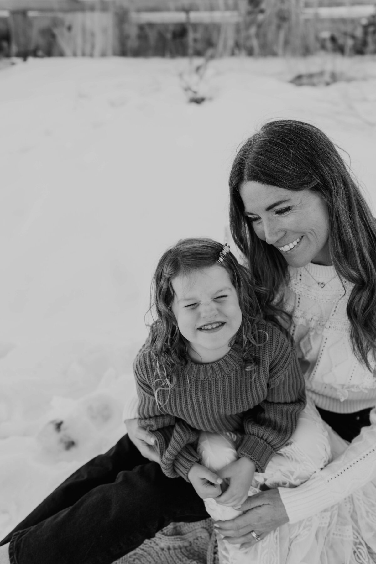 A beautiful black and white portrait of a mother adoringly, smiling at her toddler, who gives direct iContact to the portrait, while wearing an oversized brown sweater, with a beautiful snowy backdrop.