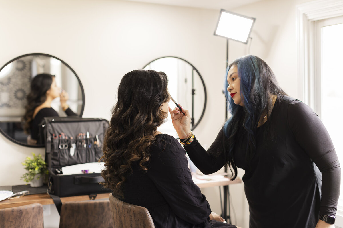 In a charming Leesburg, VA wedding venue, a makeup artist expertly applies cosmetics to a seated woman in a salon adorned with round mirrors. A professional makeup kit is open on a nearby surface, softly lit as they both dress in black, preparing for the perfect day.