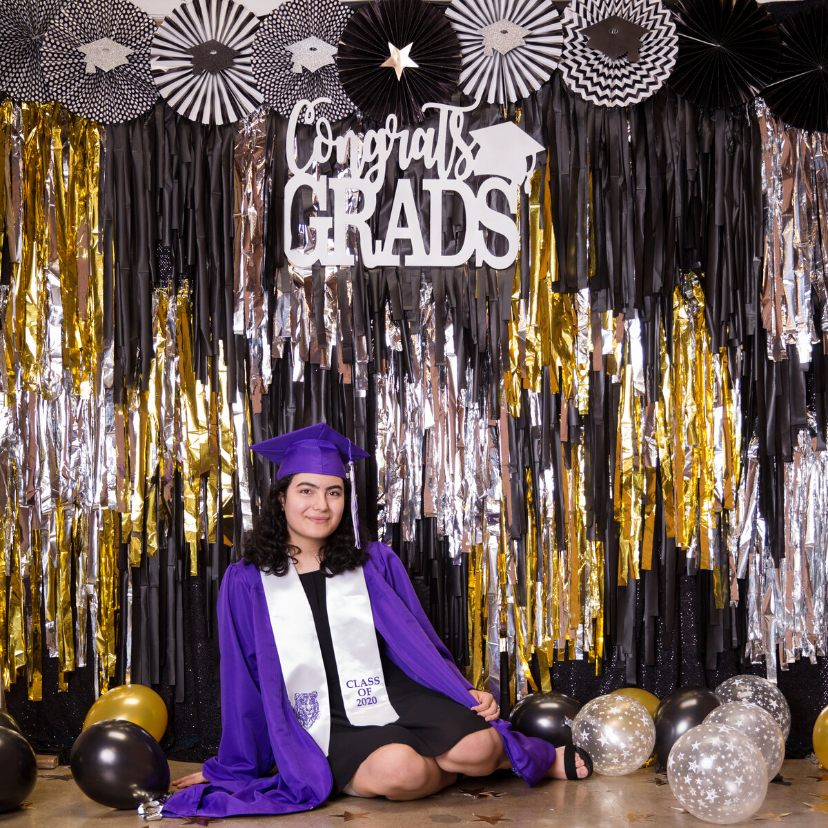 Girl in purple cap and gown posing in front of a black, gold and silver colored graduation backdrop display decorated with streamers