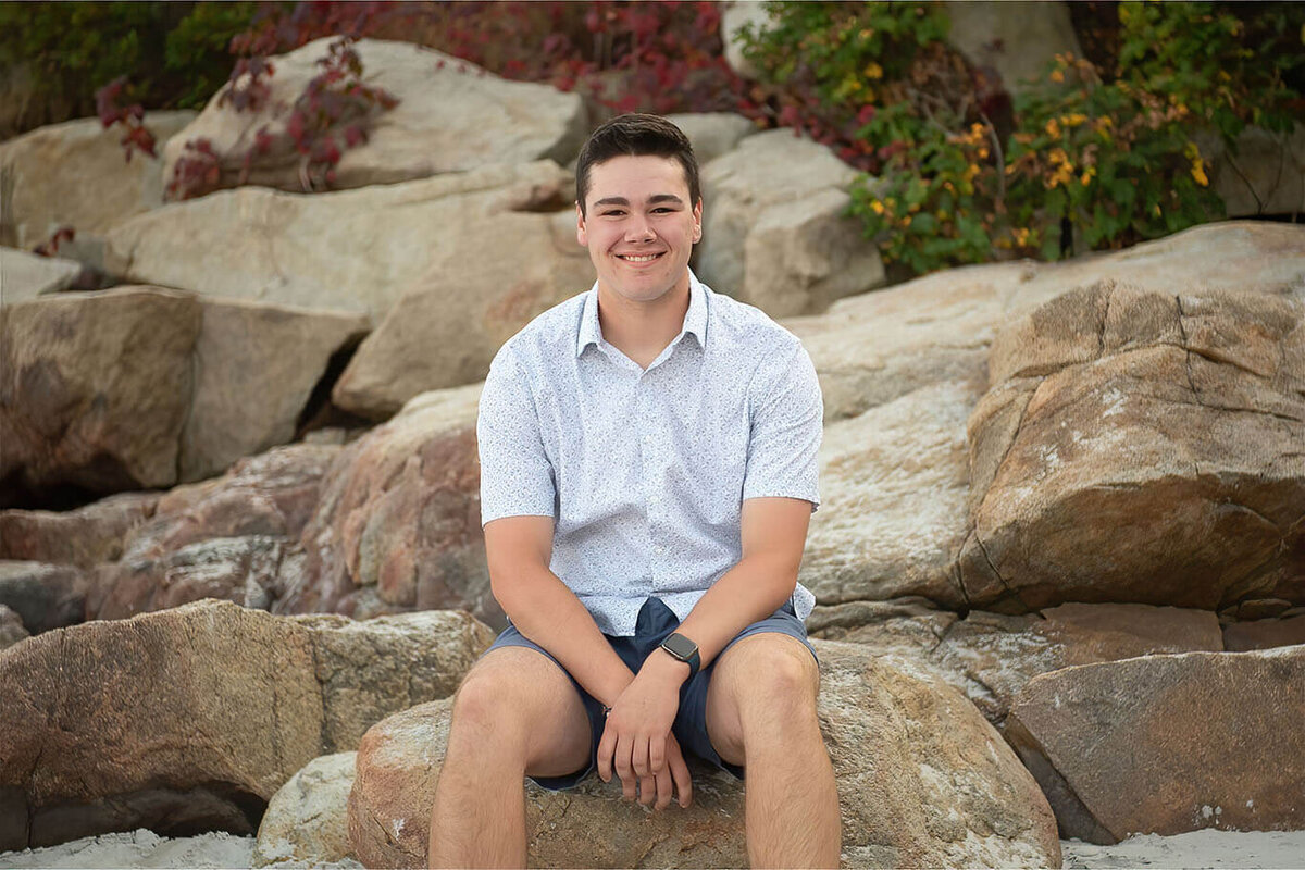 A senior boy sits on a rock at Wingaersheek Beach while smiling at the camera.