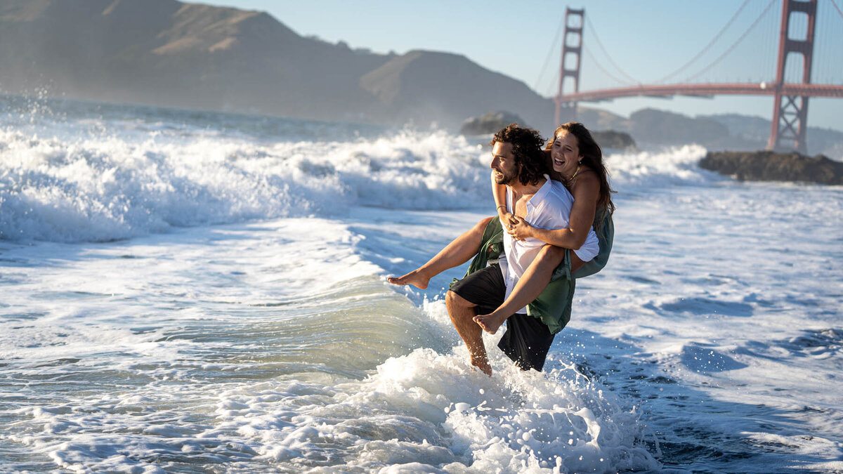 couple in a piggyback run through the surf at a beach with the golden gate bridge in the background