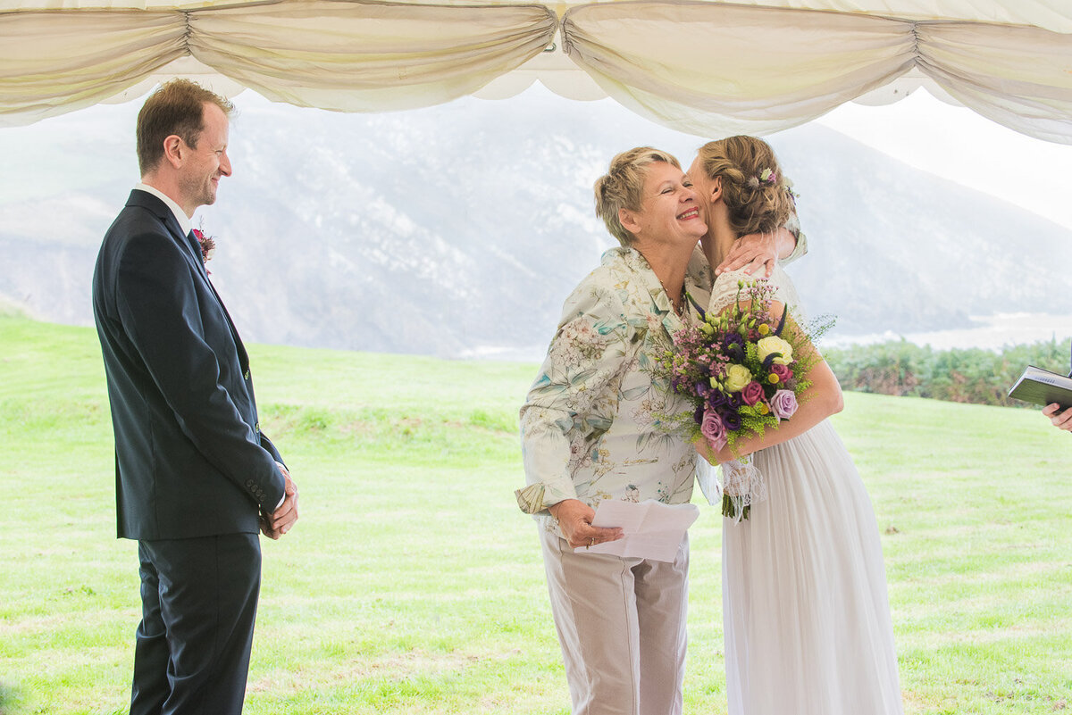 Bride wearing an empire style wedding dress hugging her Mum while holding a wild flower bouquet in a marquee overlooking the sea