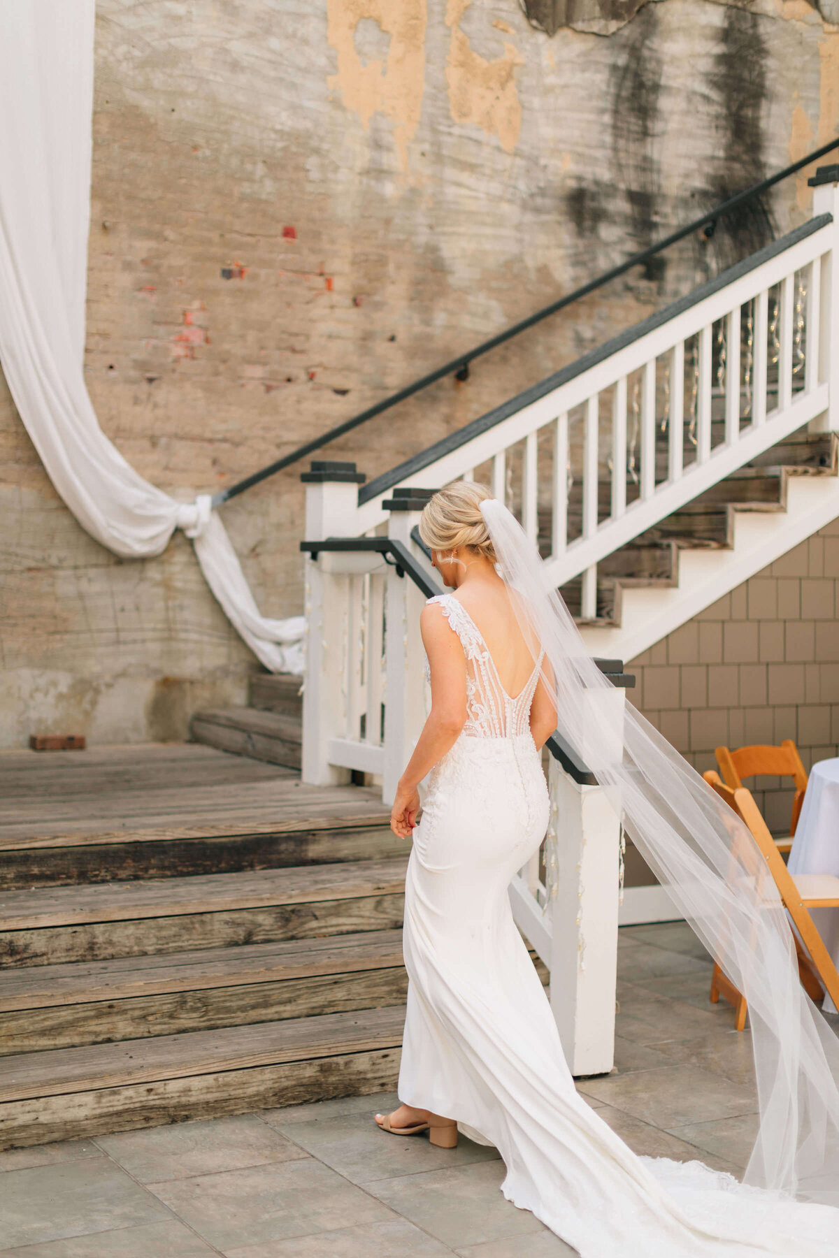 A bride in a white wedding gown with a long veil walks up wooden stairs beside a weathered brick wall draped with white fabric.