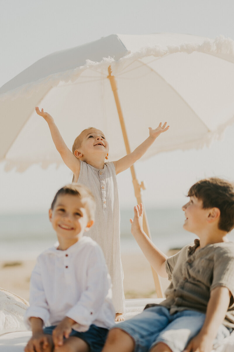 Trois petits garçons de tous âges posant et s'amusant sous une ombrelle blanche à la plage lors d'une séance photo famille en Vendée.