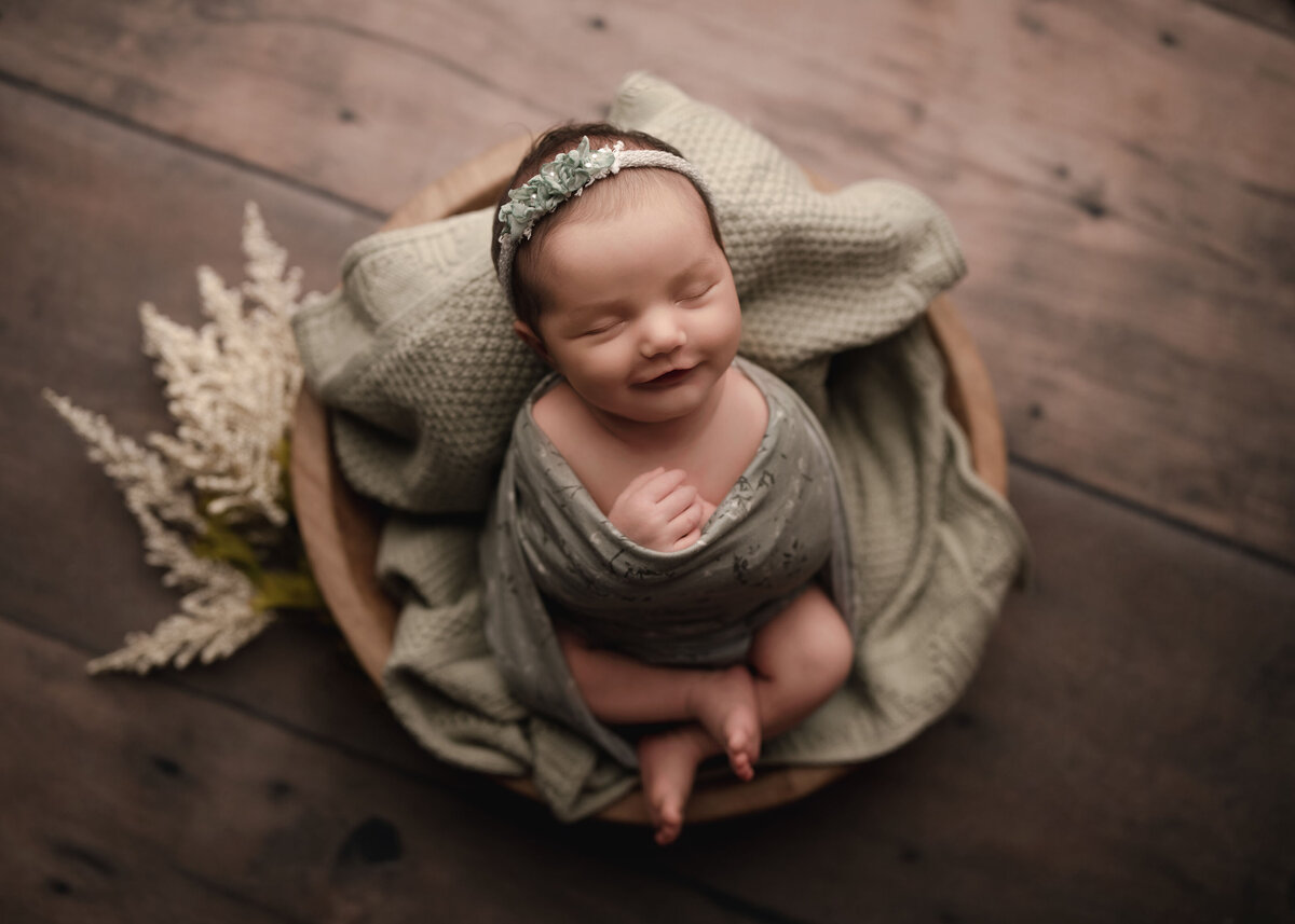 Aerial image. Newborn photoshoot. Baby girl is wrapped in a olive stretch wrap. Her legs are peeking out and folded atop of her, She is wearing a cream and green headband. Her eyes are closed with a big smile. Captured by best Menifee newborn photographer Bonny Lynn Photography