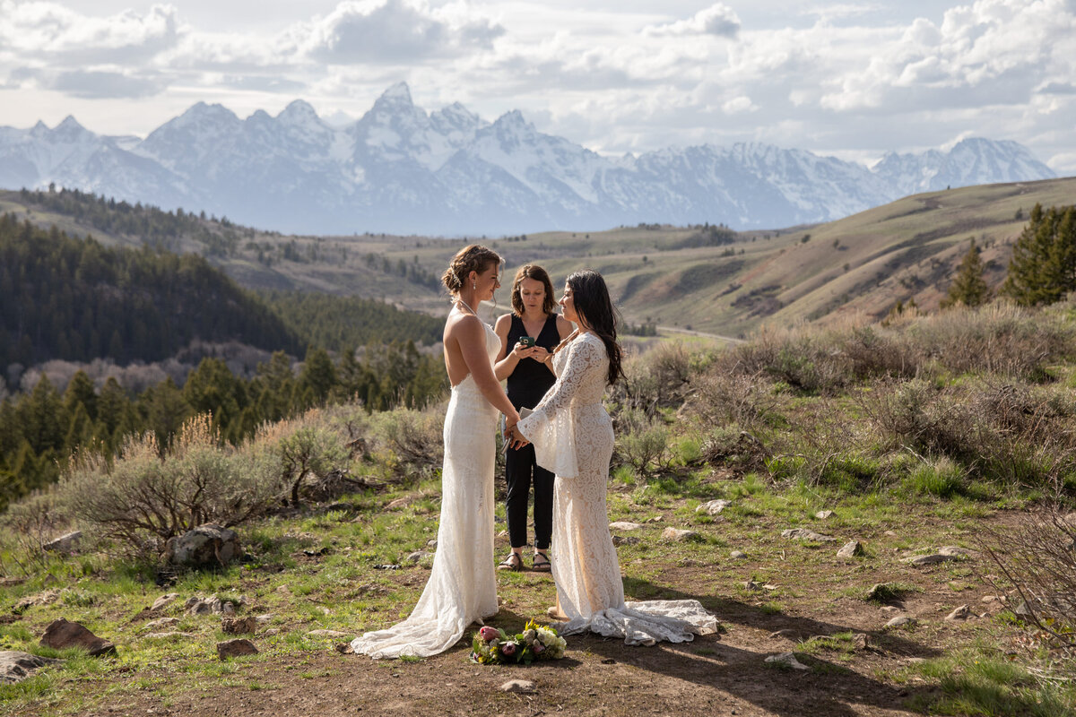 Brides stand hand in hand while the officiant performs the wedding cermony with the Tetons looming in the distance