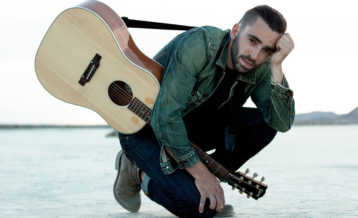 Male musician photo Michael Bernard Fitzgerald wearing jean jacket kneeling while holding pale wood guitar desert background