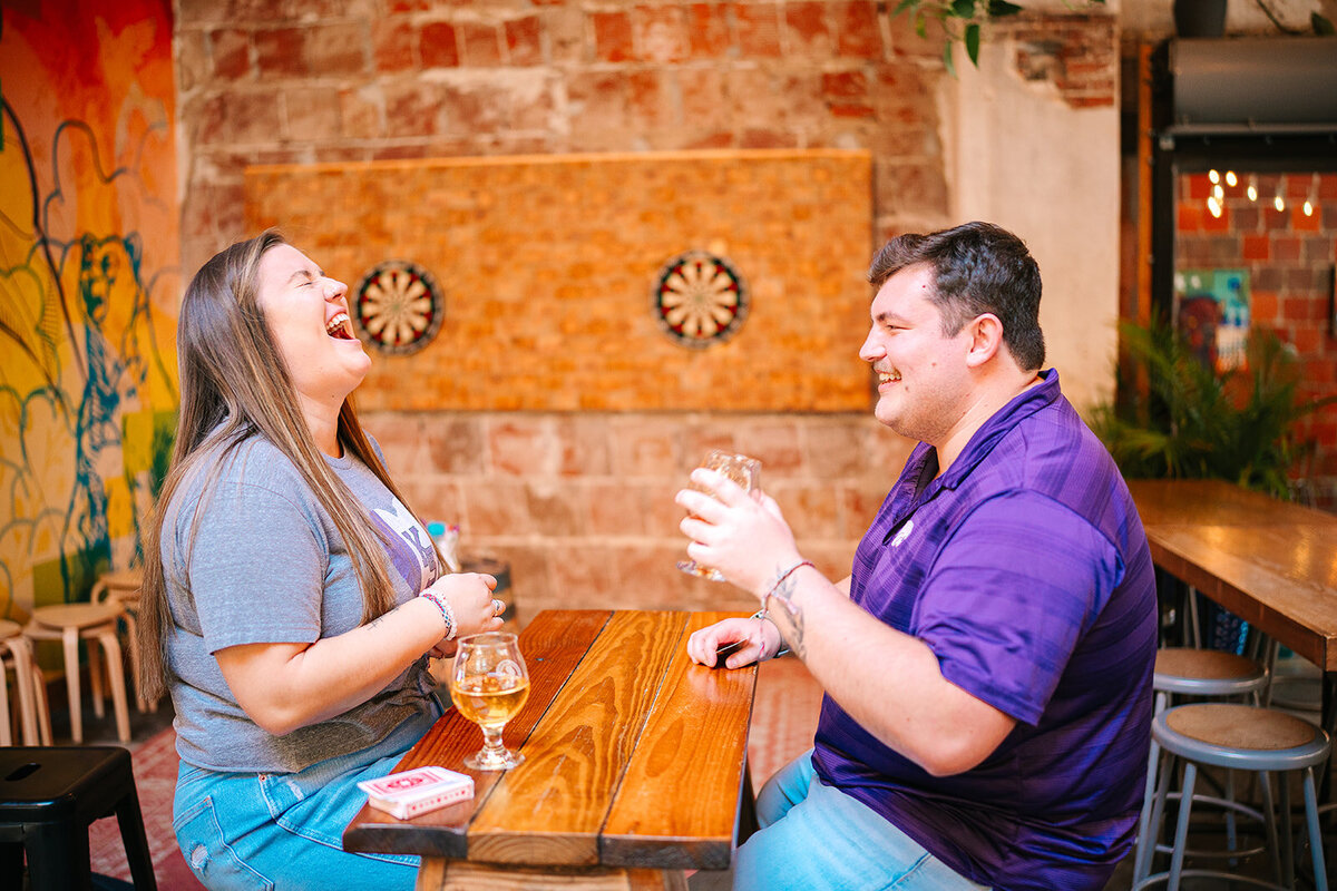 A couple laughing together while sitting on either side of a small wooden table 