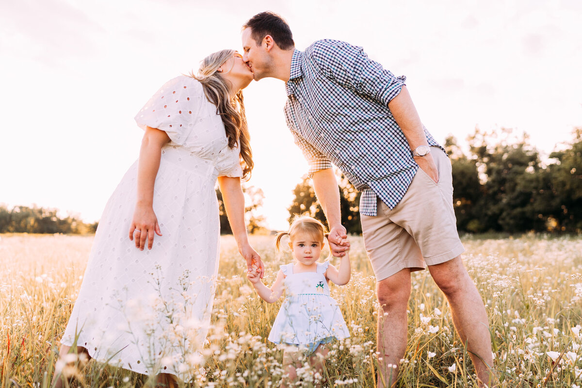 mother and father kiss while standing over 2 year old girl in wildflower field
