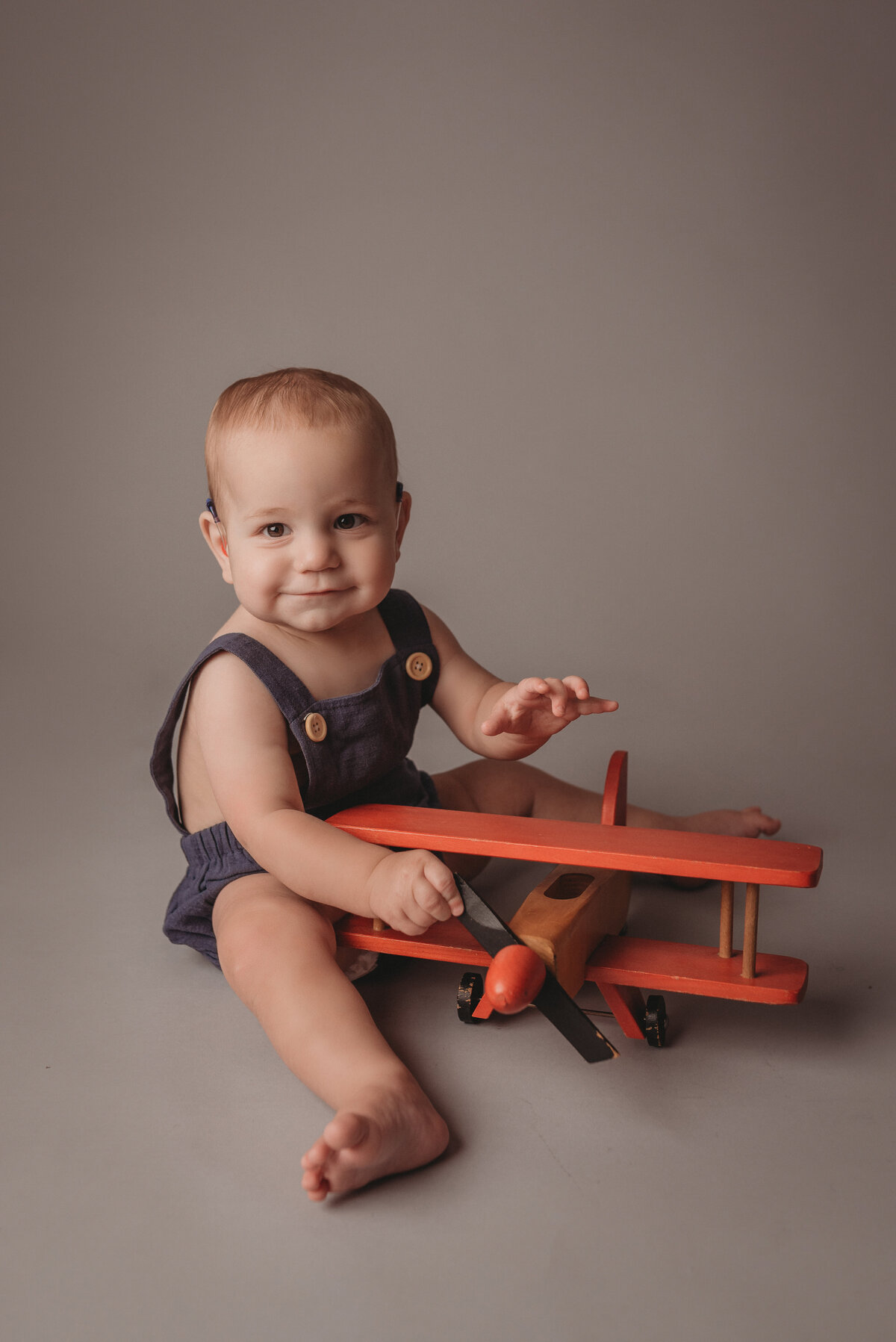 One year old boy sitting on gray backdrop wearing navy blue romper holding a wooden toy airplane