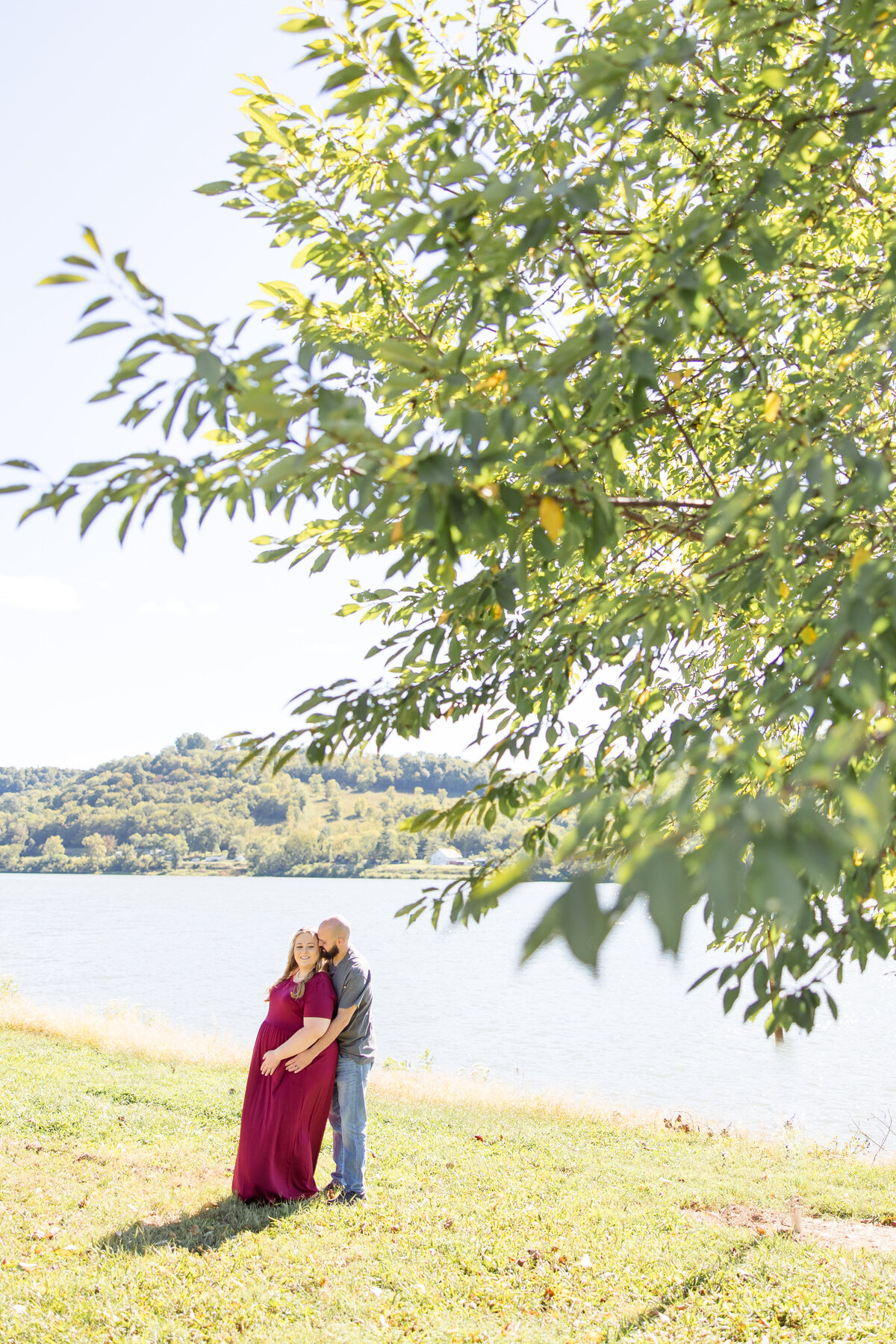 Joyful maternity photos in the bright summer sun on the Ohio River in Ripley, Ohio. Husband and wife snuggled back to front cuddling in close.