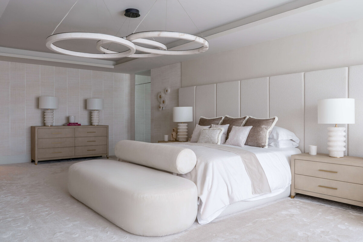 A spacious primary bathroom with a  bright, airy feel. The large, white bed is decorated with neutral accent pillows, with light wood side tables at each side. There is a large white footboard bench, and above the bed is a large, contemporary light fixture. composed of three circles. On one side of the bed is a light dresser that matches the side tables.