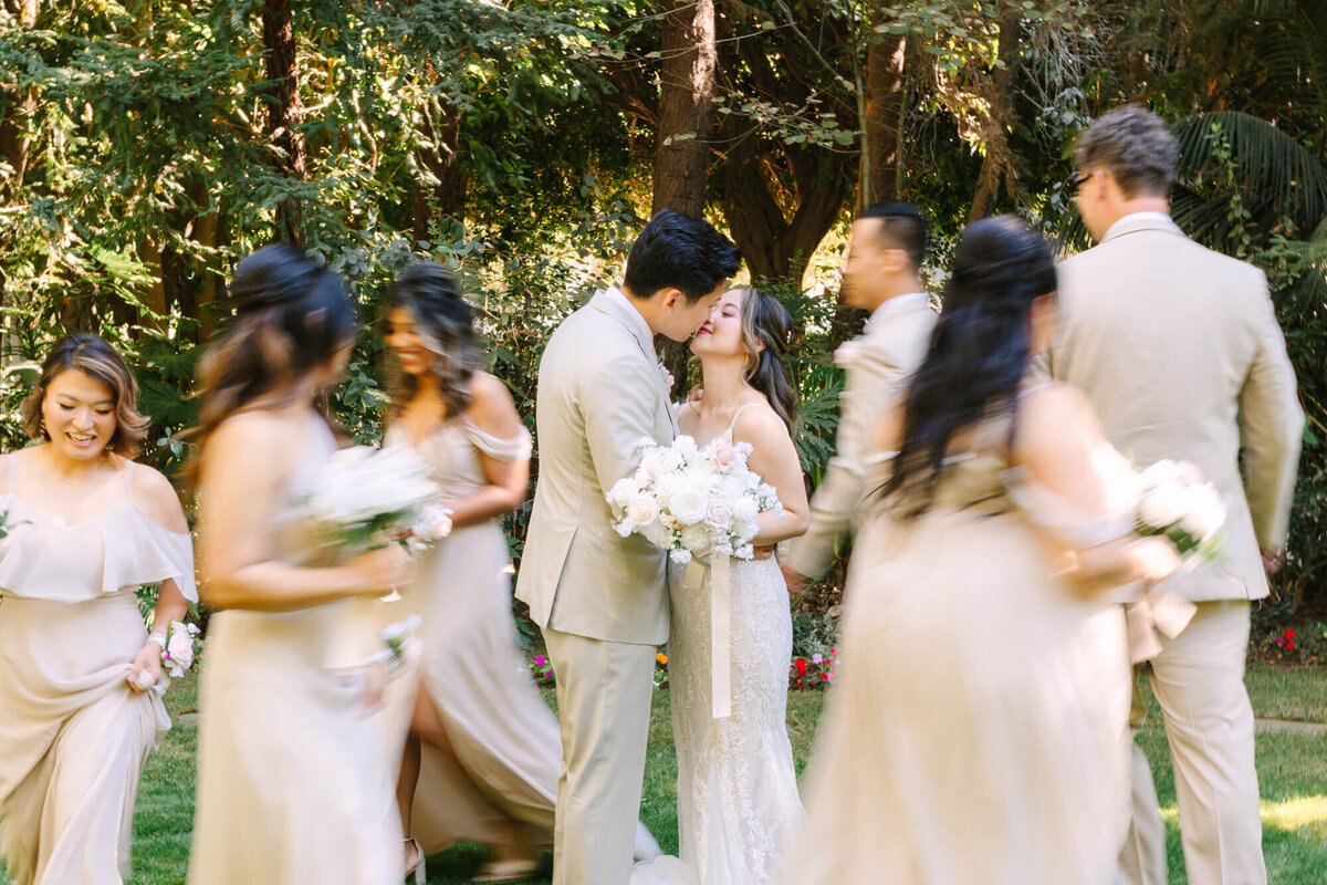 Bride and groom kiss in a garden, surrounded by bridal party in motion. The couple embraces, holding a white bouquet.