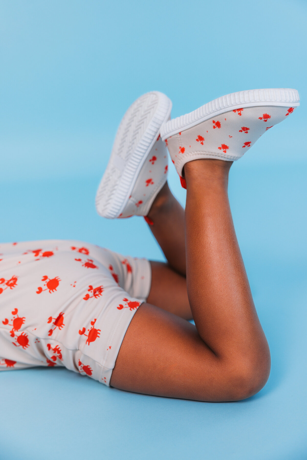 Child wearing Swim Essentials UV shorts and matching water shoes with a playful red crab print, lying on a blue surface with legs playfully raised.