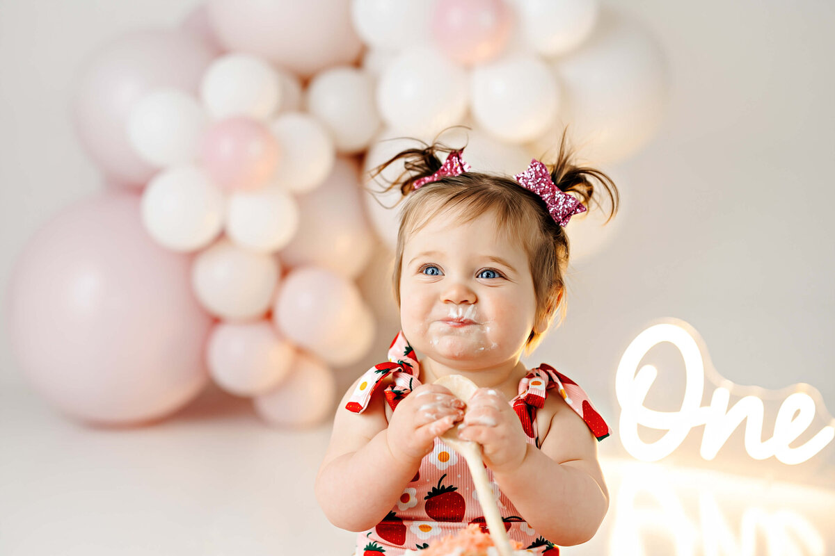 A baby with pigtails, wearing a strawberry-patterned dress, surrounded by pink and white balloons. The child has cake on their face and holds a piece of cake. A lit sign saying "One" is visible beside them.