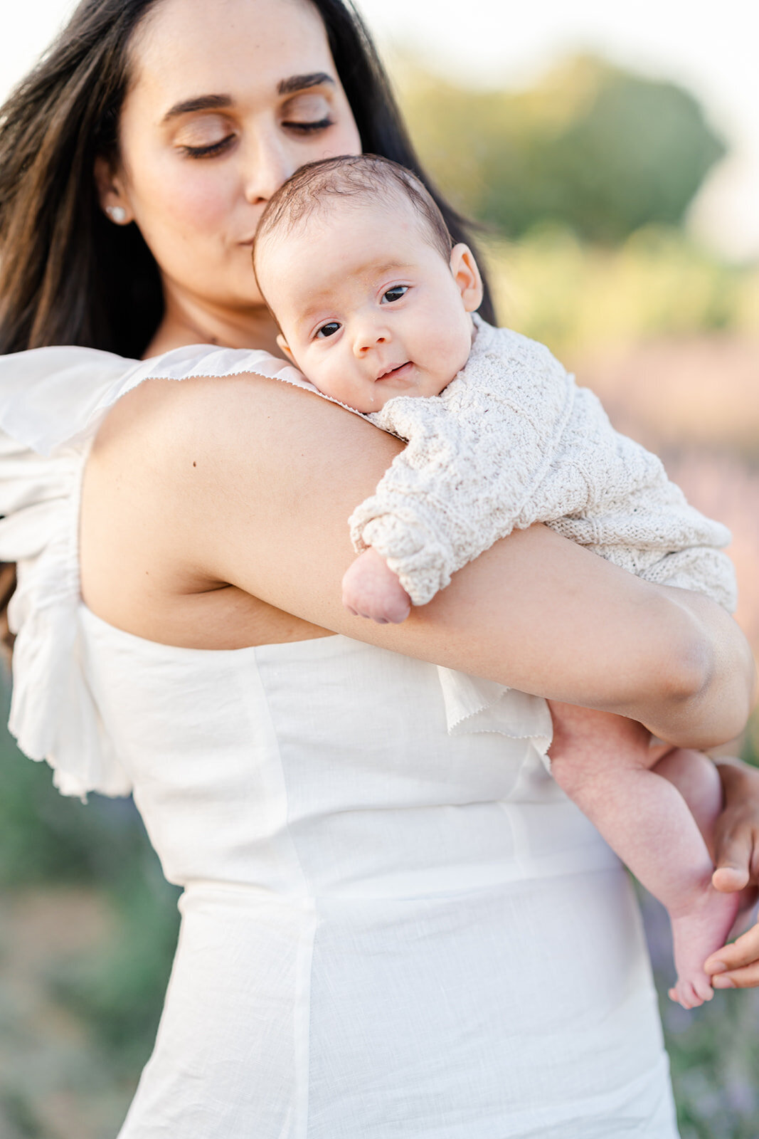A mother wearing a white gown kisses the head of her newborn baby as the baby hugs her shoulder photographed by Bay Area Photographer, Light Livin Photography.