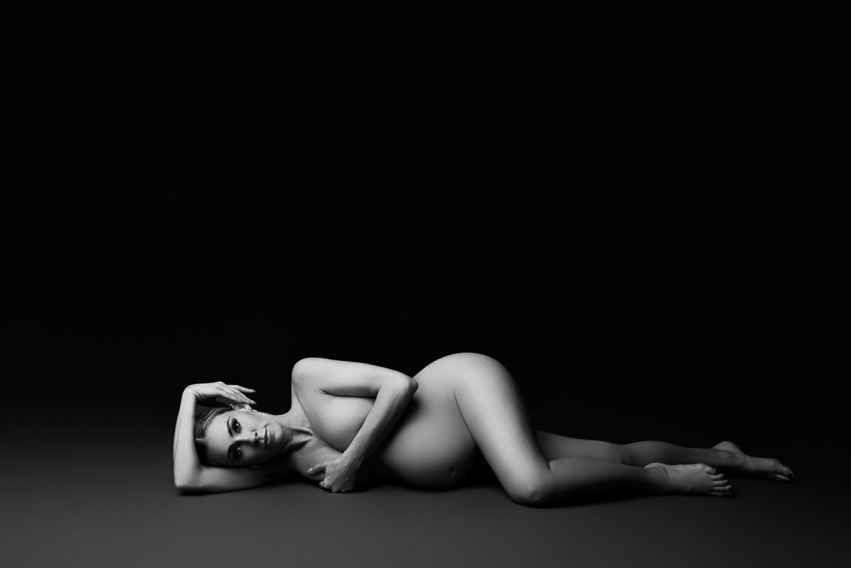Black and white image of a woman posing for a New Jersey fine art maternity photoshoot. The woman lies bare on the ground, facing the camera. One arm is under her head, with her hand artfully resting atop her head, while the other arm provides modest coverage to her breasts. Her top leg is gracefully bent in front of her. This captivating image was skillfully captured by Katie Marshall, recognized as the best maternity photographer in New Jersey.