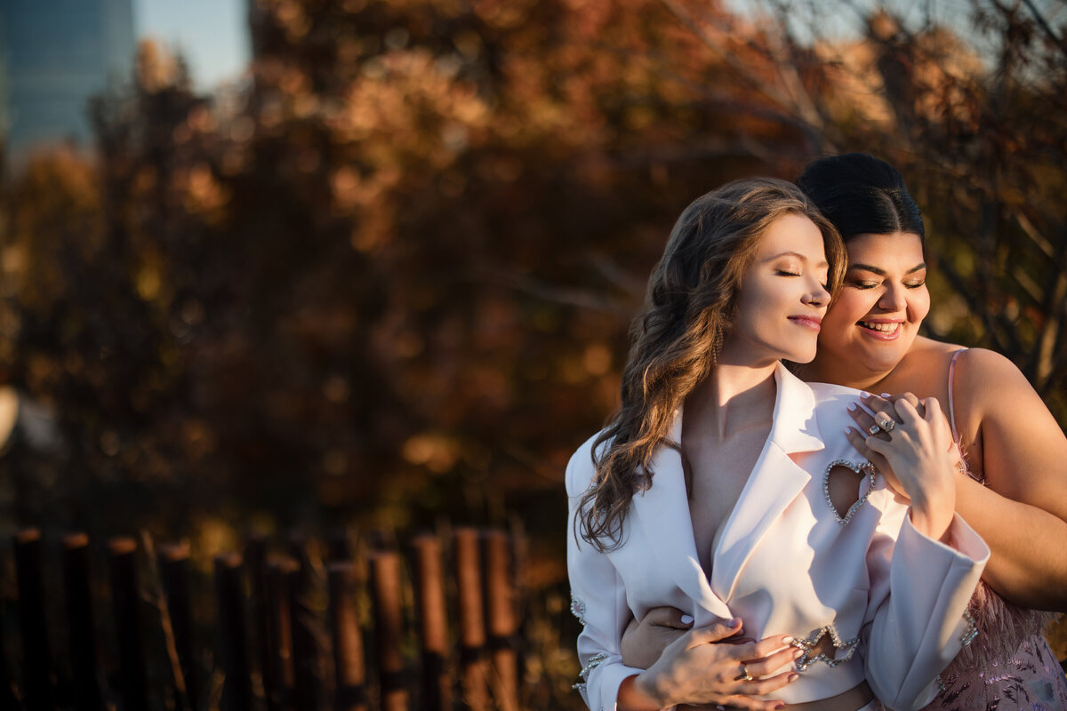 Two brides stand closely, one in a white blazer and the other in a pink dress, smiling and embracing in a sunlit outdoor setting with blurred autumn foliage in the background.  This is at a Little Island NYC sunset engagement session