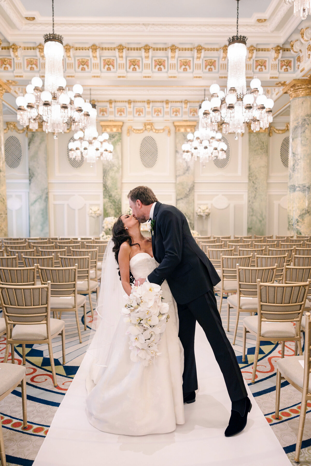 A bride and groom share a kiss in an elegant ballroom lined with empty chairs. The bride holds a bouquet of white flowers and wears a strapless gown, while the groom is dressed in a dark suit. Chandeliers and ornate columns adorn the room.