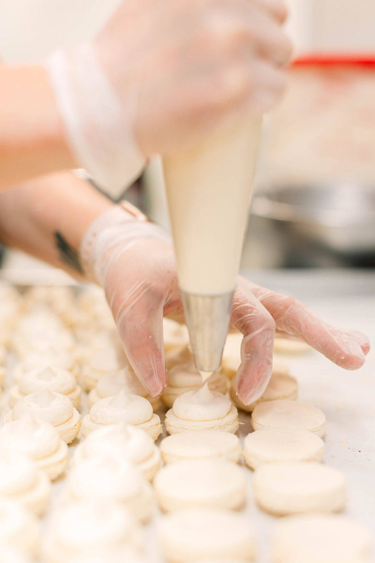 A baker wearing gloves carefully pipes creamy filling onto freshly baked macaron shells, assembling delicate pastries in a professional kitchen.